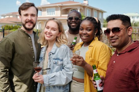 Foto de Feliz joven amigos interculturales con bebidas alcohólicas mirando a la cámara mientras disfruta de la reunión al aire libre contra la casa de campo - Imagen libre de derechos