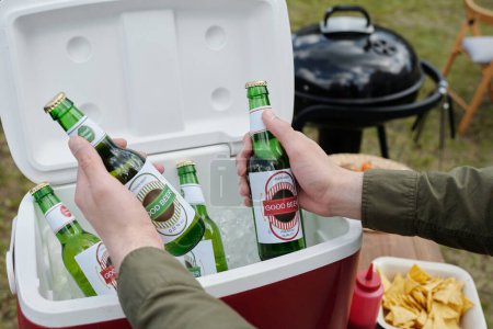 Photo for Hands of young man holding two bottles of beer over box prepared for guests and backyard party against barbecue grill and potato chips in bowl - Royalty Free Image
