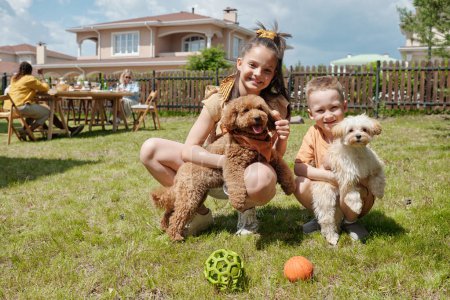Téléchargez les photos : Deux enfants joyeux mignons jouant avec leurs animaux de compagnie de chien sur l'herbe verte de l'arrière-cour de leur maison de campagne le jour ensoleillé d'été ou week-end - en image libre de droit