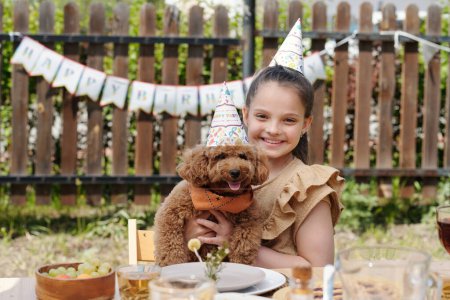 Téléchargez les photos : Bonne petite fille dans le chapeau d'anniversaire tenant chien animal de compagnie mignon tout en étant assis à table servie pendant le dîner en plein air ou la fête à l'arrière-cour de la maison de campagne - en image libre de droit