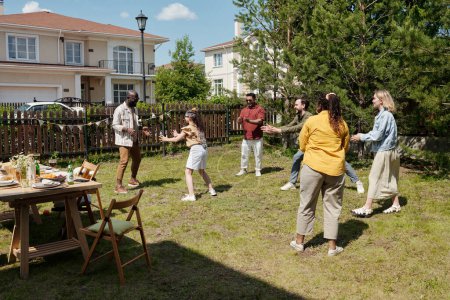 Foto de Grupo de hombres, mujeres y niños interculturales felices jugando al escondite en la hierba verde del patio trasero de su casa de campo el fin de semana - Imagen libre de derechos