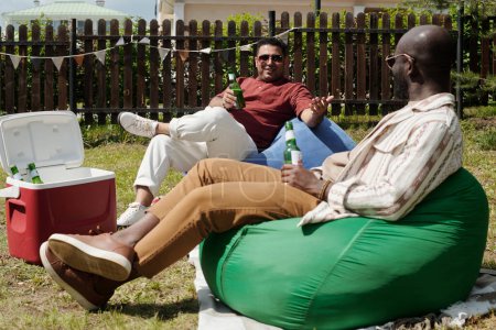 Foto de Dos jóvenes hombres interculturales con botellas de cerveza sentados en sillas de bolsa y charlando durante la fiesta al aire libre en el campo el fin de semana - Imagen libre de derechos