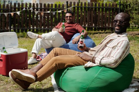 Foto de Dos jóvenes y felices hombres interculturales con botellas de cerveza sentados en sillas de bolsa y mirando a la cámara durante la fiesta al aire libre el fin de semana - Imagen libre de derechos