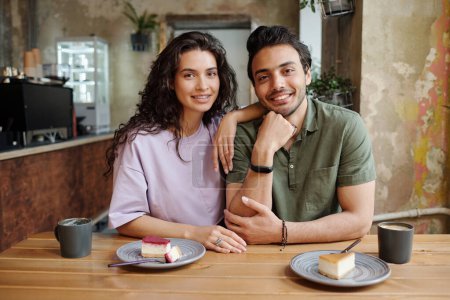 Foto de Joven pareja alegre mirando a la cámara mientras está sentado junto a la mesa y teniendo sabrosos pasteles de queso y capuchino durante la cita romántica en la cafetería - Imagen libre de derechos