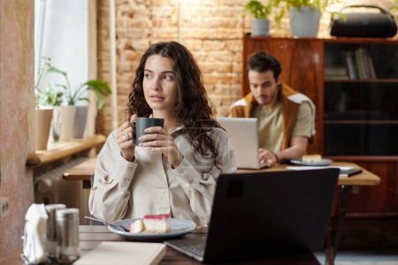 Téléchargez les photos : Jeune indépendante reposante en tenue décontractée assise à table devant un ordinateur portable dans un café et regardant à travers la fenêtre par une tasse de café - en image libre de droit