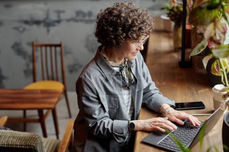 Photo for Serious middle aged businesswoman or freelancer with laptop sitting in front of window in cafe, typing on keyboard and looking at screen - Royalty Free Image