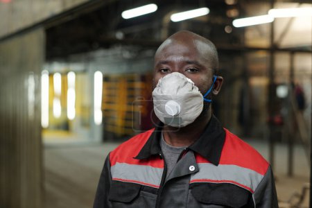 Foto de Joven hombre afroamericano en ropa de trabajo y respirador protector de pie delante de la cámara contra el interior del almacén - Imagen libre de derechos