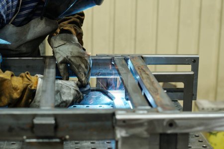Téléchargez les photos : Mains gantées de travailleurs masculins de l'usine industrielle produisant d'énormes machines se penchant sur l'établi et soudant des pièces métalliques - en image libre de droit