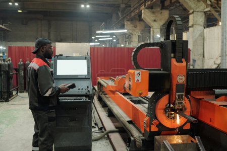 Foto de Joven trabajador masculino de planta industrial en pie por terminal de control mientras comprueba el trabajo de la máquina de producción en taller - Imagen libre de derechos