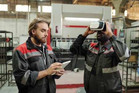 Photo for Young male engineer in workwear scrolling in tablet while standing by colleague in vr headset making presentation of new equipment - Royalty Free Image