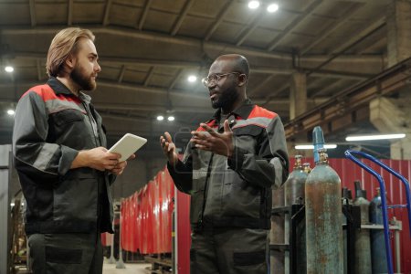 Téléchargez les photos : Jeune ingénieur masculin sérieux regardant son collègue afro-américain décrire les caractéristiques de nouveaux équipements industriels - en image libre de droit