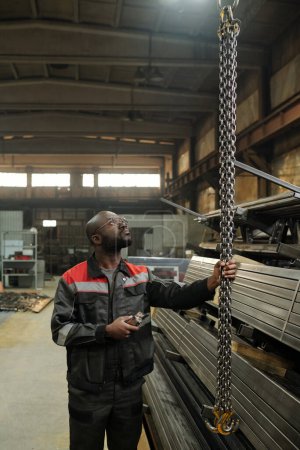 Foto de Joven ingeniero afroamericano mirando hacia arriba mientras tira de un montón de cadenas frente a pilas de repuestos metálicos para máquinas industriales - Imagen libre de derechos