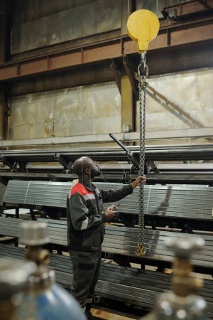 Foto de Joven hombre negro tirando de racimo de cadenas metálicas mientras está de pie delante de pilas de materiales para la construcción de máquinas industriales - Imagen libre de derechos