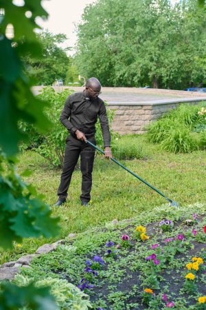 Photo for Young African American pastor in black pants and shirt with clerical collar working with rake in the garden or backyard by church building - Royalty Free Image