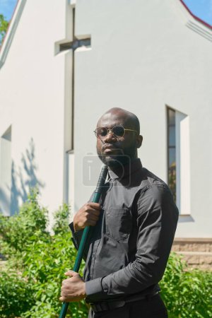 Photo for Young serious pastor of African American ethnicity holding worktool while standing in church garden against white building and looking at camera - Royalty Free Image