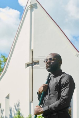 Téléchargez les photos : Jeune pasteur sérieux de l'église luthérienne en lunettes et chemise noire avec col clérical tenant râteau contre bâtiment blanc avec croix - en image libre de droit
