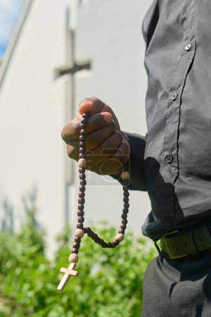 Téléchargez les photos : Main de jeune prêtre afro-américain avec des perles de chapelet en bois disant la prière ou pratiquant la prière silencieuse dans le jardin par la construction de l'église - en image libre de droit