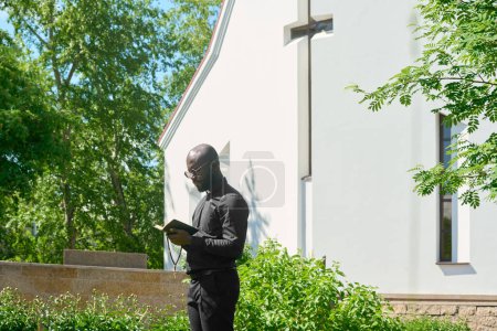 Téléchargez les photos : Vue latérale du jeune prédicateur en vêtements noirs regardant dans la Sainte Bible ouverte tout en lisant les versets de l'un des évangiles canoniques à l'extérieur - en image libre de droit