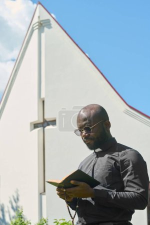 Foto de Hombre afroamericano serio con la Santa Biblia abierta y cuentas de rosario de madera leyendo versículos del Evangelio contra la construcción de la iglesia - Imagen libre de derechos