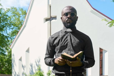 Photo for Young black man in shirt with clerical collar standing with open Bible in front of camera while preaching in front of church building - Royalty Free Image
