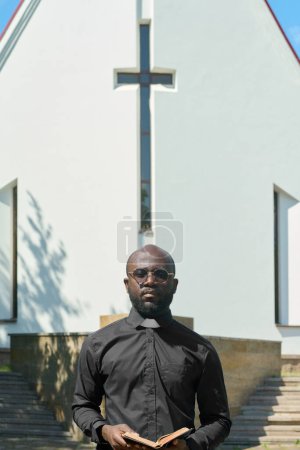 Photo for Serious black man with open Gospel in hands looking at camera against white building of Lutheran church with cross on the front part - Royalty Free Image
