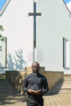 Photo for Young serious priest with open Bible evangelizing audience or telling sermon while standing in front of building of Catholic church - Royalty Free Image