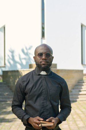 Foto de Joven sacerdote serio con camisa negra con cuello clerical sosteniendo la Sagrada Biblia mientras reza o pronuncia un sermón frente al edificio de la iglesia - Imagen libre de derechos