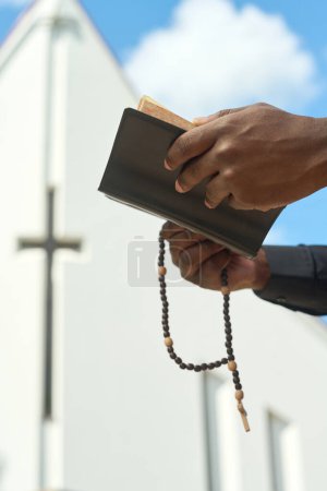 Photo for Hands of black man holding open Holy Bible in leather cover and wooden rosary beads while reading Gospel and saying prayers to Lord - Royalty Free Image