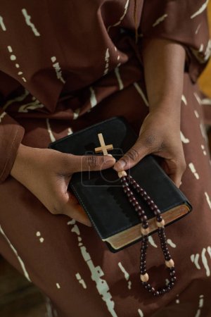Foto de Manos de mujer negra joven en vestido marrón sosteniendo la Biblia y rosarios con una pequeña cruz de madera mientras asisten al servicio de la iglesia - Imagen libre de derechos