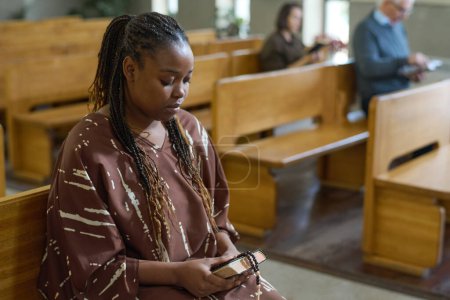 Foto de Joven mujer afroamericana sosteniendo las cuentas de la Biblia y del rosario mientras está sentada en el banco en la iglesia evangélica durante el sermón - Imagen libre de derechos