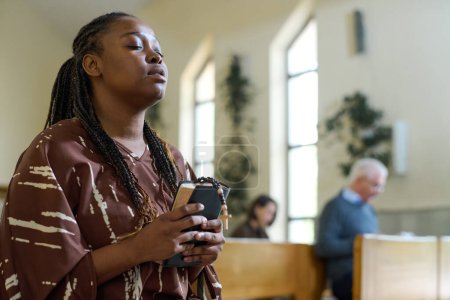 Foto de Joven mujer negra en vestido casual manteniendo los ojos cerrados durante la oración silenciosa en la iglesia mientras sostiene la Santa Biblia y las cuentas del rosario - Imagen libre de derechos