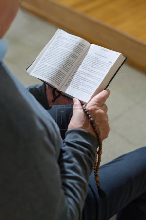 Photo for Hands of senior man with open Bible or Gospel and rosary beads reading verses while sitting in church during sermon or service - Royalty Free Image