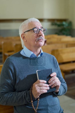 Photo for Aged serious man with grey hair holding Holy Bible and wooden rosary beads by chest while concentrating on pray while sitting in church - Royalty Free Image