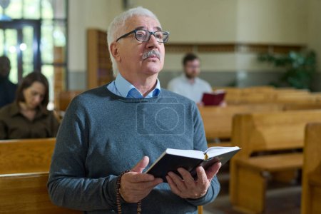 Photo for Mature man with grey hair holding open Bible while sitting on bench during church service, looking at preacher and listening to sermon - Royalty Free Image