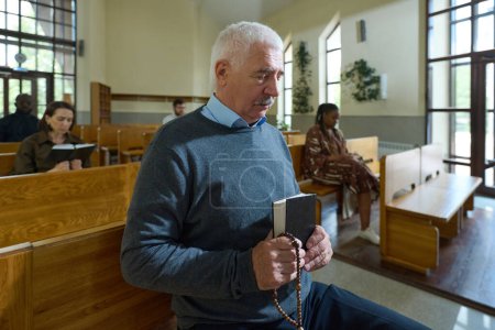 Photo for Mature man with Holy Bible and rosary beads in hands saying prayers to himself while sitting on bench during church service - Royalty Free Image