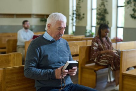 Téléchargez les photos : Homme aux cheveux gris sérieux en tenue décontractée tenant devant lui des perles de la Bible et du chapelet pendant la prière silencieuse contre une femme noire à l'église - en image libre de droit