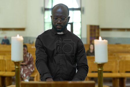 Foto de Sacerdote afroamericano de camisa negra con cuello clerical de pie junto al púlpito de madera entre dos velas encendidas durante el servicio de la iglesia - Imagen libre de derechos