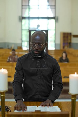 Foto de Joven afroamericano de camisa negra con cuello clerical leyendo uno de los cuatro Evangelios o algún otro libro de la Sagrada Biblia durante la liturgia - Imagen libre de derechos