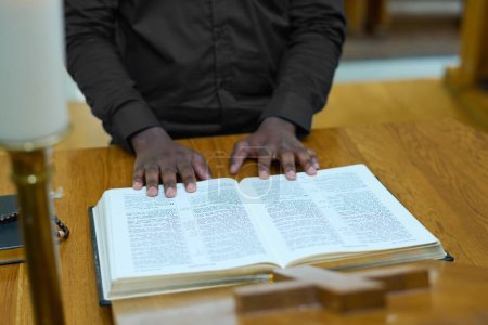 Hands of young black man on pages of open Holy Bible standing by wooden pulpit during sermon in Catholic church
