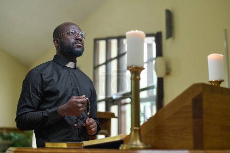 Photo for African American priest in black casualwear holding rosary beads during pray while standing by pulpit with two burning candles - Royalty Free Image