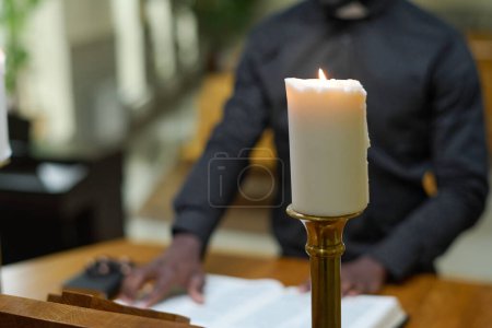 Foto de Vela blanca encendida en el fondo del sacerdote leyendo versículos de la Santa Biblia mientras está de pie junto al púlpito y diciendo sermón durante el servicio a la iglesia - Imagen libre de derechos