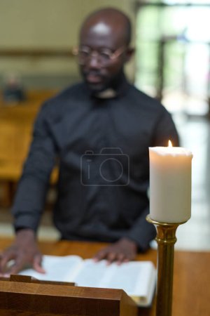 Téléchargez les photos : Bougie blanche allumée contre un jeune pasteur afro-américain lisant la Sainte Bible en se tenant debout à la chaire devant des paroissiens - en image libre de droit