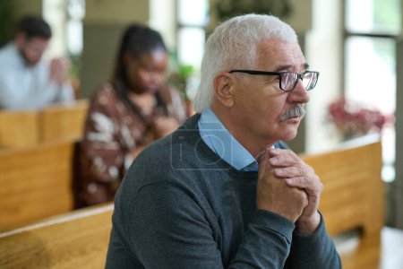 Photo for Mature male parishioner with grey hair praying with his hands put together while sitting on bench against other people - Royalty Free Image
