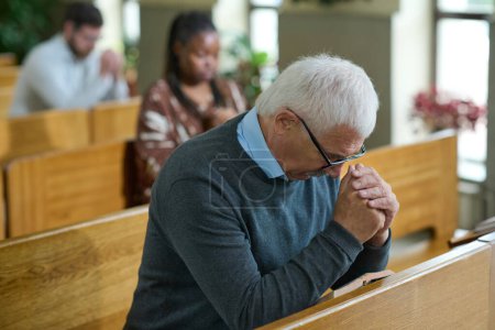 Photo for Mature man inclining his head and bending over his hands put together during silent pray after sermon or before communion in church - Royalty Free Image