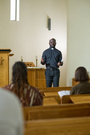 Photo for Young black man in shirt with clerical collar and pants preaching in front of group of intercultural parishioners sitting on benches in church - Royalty Free Image