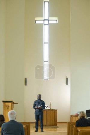 Téléchargez les photos : Prêtre afro-américain en tenue décontractée noire faisant sermon pour les paroissiens tout en se tenant contre la chaire et le mur avec croix - en image libre de droit