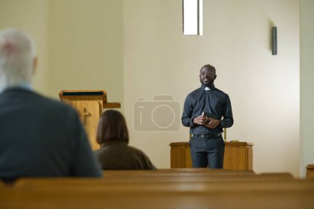 Photo for Young black man in priest apparel with clerical collar standing in front of parishioners and explaining verses from Holy Bible during sermon - Royalty Free Image