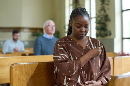 Foto de Joven mujer negra con los ojos cerrados cruzándose durante la oración silenciosa mientras se sienta en el banco contra los feligreses en la iglesia - Imagen libre de derechos