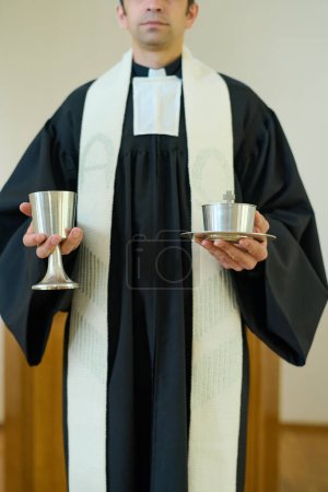 Photo for Clergyman in cassock with clerical collar holding cups with communion stuff while praying before oblation and waiting for parishioners - Royalty Free Image