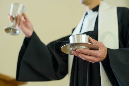 Photo for Catholic priest holding cups with wine and unleavened bread symbolizing flesh and blood of Savior prepared for communion of parishioners - Royalty Free Image